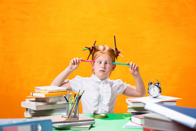 Menina de, aproximadamente, 10 anos, em um fundo amarelo, sentada em frente a uma mesa verde. Ela está desfocada dos estudos, começou a brincar com os materiais, fazer caretas e colocar vários lápis e canetas no cabelo e no rosto. Na mesa tem diversos objetos como livros, canetas, cadernos, relógio, porta lápis e tesoura.