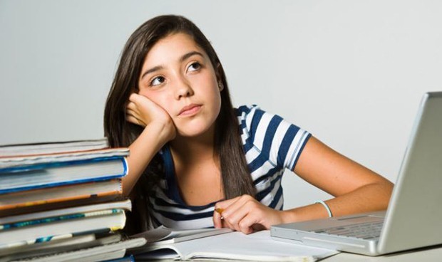 Garota de, aproximadamente, 15 anos, em um fundo cinza, sentada e apoiando seus braços em uma mesa de cor semelhante, a qual está cheia de livros, cadernos e um notebook. Ela está com um olhar distante, sem conseguir ter foco e atenção nos estudos.