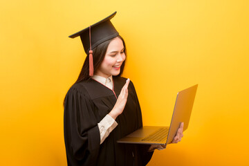 Jovem de, aproximadamente, 17 anos, em um fundo amarelo e vestindo um capelo e uma beca (roupa usada em formaturas). Ela está segurando um notebook enquanto faz uma chamada de vídeo e está completando o Ensino Médio através do método EAD.